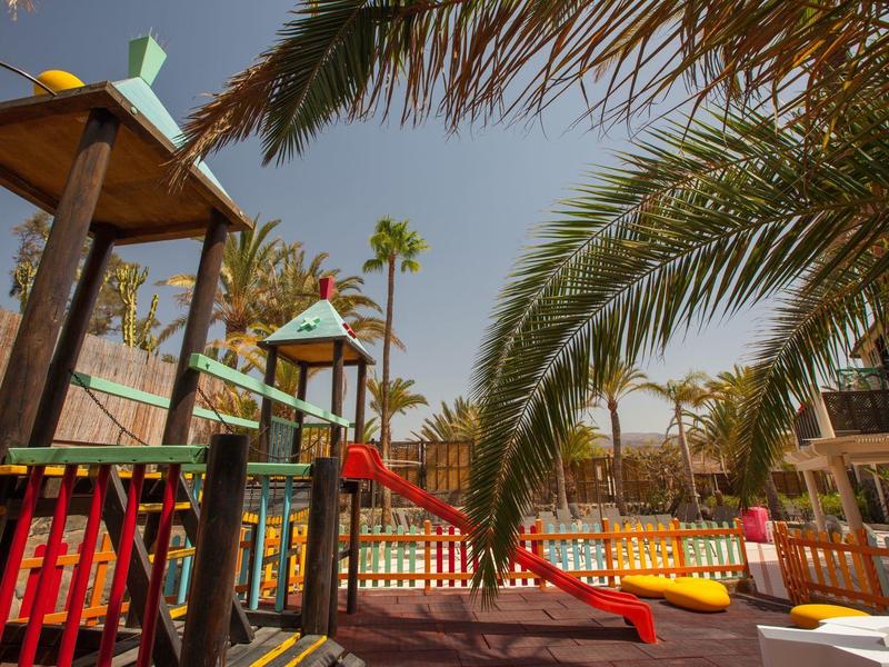 Colorful playground with slides and climbing frames under palm trees in a sunny hotel garden.