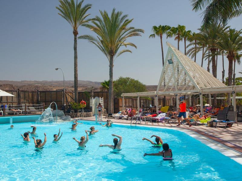 People swimming outdoors in a pool with a pavilion and palm trees nearby.