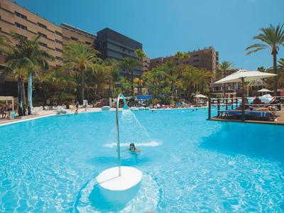 Duży basen hotelowy z leżakami i palmami w słoneczną pogodę.