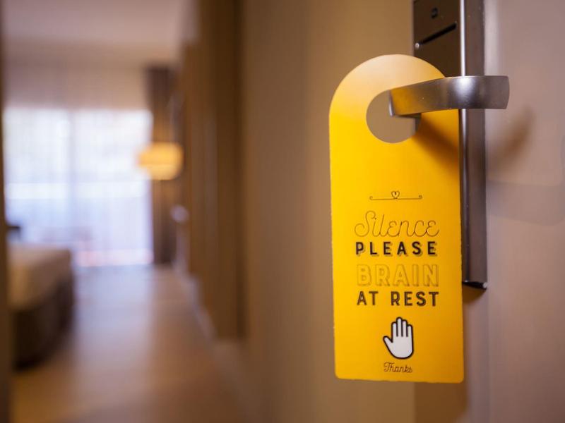 Cartel amarillo colgado en puerta de hotel pidiendo silencio, con cama y lámpara desenfocadas al fondo.