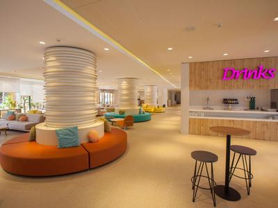 Een moderne hotel-lounge met ronde zitplaatsen, zuilen en een bar met een bord 'Drinks'.
