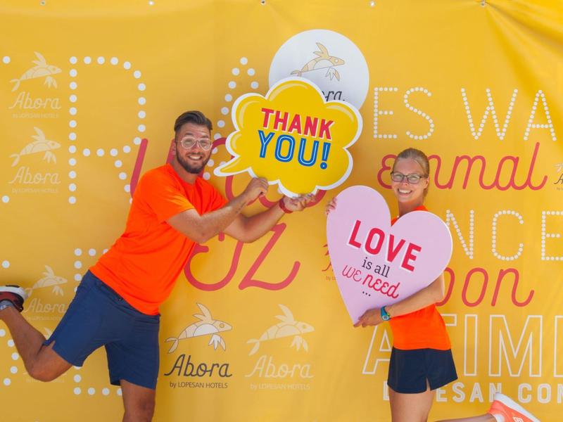 Dos personas alegres con camisetas naranjas posan frente a fondo amarillo con letreros de gracias y amor.