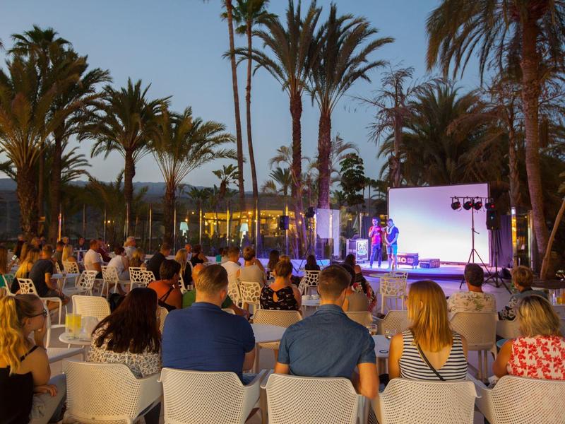 Evento nocturno con público y escenario bajo palmeras en un jardín de hotel.