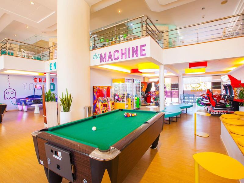 Área de juegos bien iluminada con mesa de billar, asientos y máquinas arcade en un hotel.