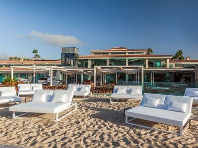 Sonnenliegen und Sofas stehen auf Sand vor modernem Hotelgebäude unter blauem Himmel.