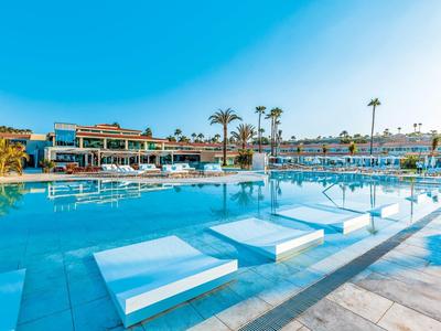 Large pool with sunbeds submerged in water at a sunny resort with palm trees and clear blue sky.