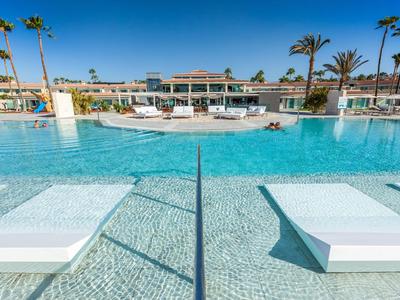 Grande piscina dell'hotel con lettini nell'acqua e palme sullo sfondo sotto un cielo blu.