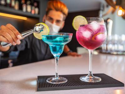 Bartender pouring a blue cocktail into a martini glass next to a pink cocktail with lime on a bar counter.