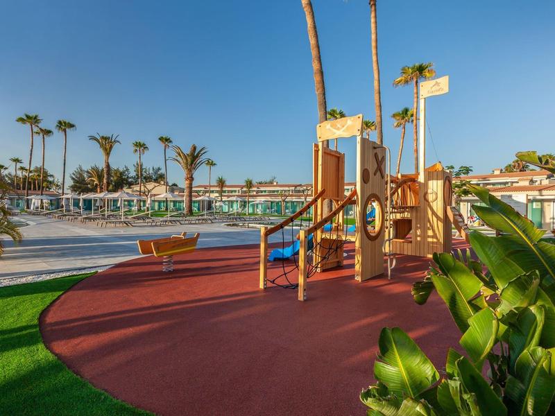 Children's playground with climbing frame near large outdoor swimming pool under palm trees.