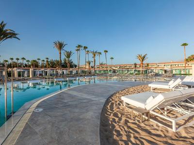 Piscina all'aperto dell'hotel con lettini e palme sotto un cielo azzurro.