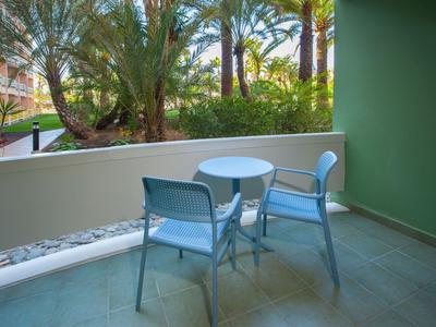 Twee stoelen en een ronde tafel op een balkon met uitzicht op palmbomen en groen.