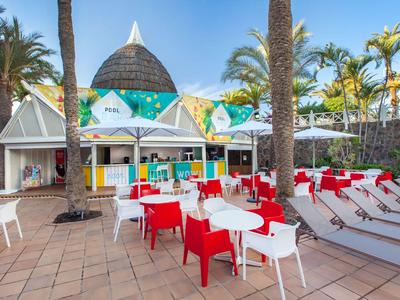 Kleurrijk terras met rode en witte stoelen, tafels en witte ligbedden onder palmbomen in een vakantieresort.