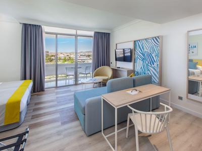 Bright hotel room with balcony, blue sofa, desk, and sea view.