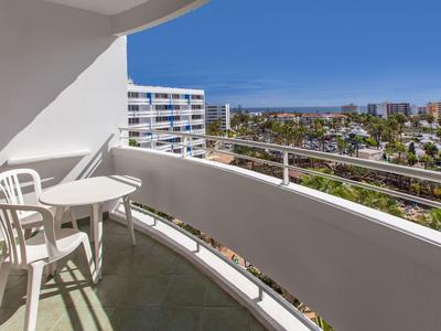 Balcony with white railing, two white chairs, and small round table overlooking buildings and palm trees.