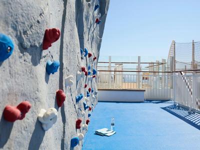 Climbing wall with colorful holds on a blue floor with railing and clear sky in the background.