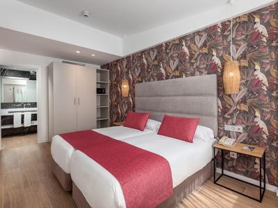 Modern hotel room with double bed, red bedding, floral accent wall, and adjoining bathroom.