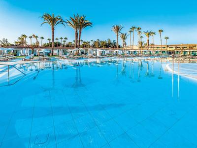 Large pool with clear blue water, surrounded by palm trees and lounge chairs under a clear sky.