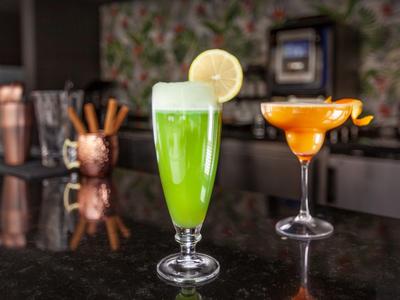 Dos cócteles coloridos en la barra con una bebida verde y un cóctel naranja