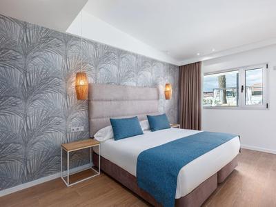 Modernes Hotelzimmer mit großem Bett, blauer Bettdecke, Akzentwand und Fenster mit Blick nach draußen.