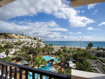 Vue du balcon d'un hôtel avec piscine, palmiers et mer sous un ciel nuageux.