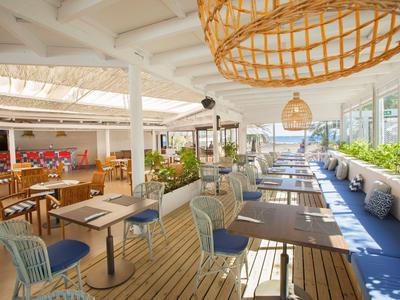 Restaurante moderno y luminoso con suelo de madera, sillas de mimbre y grandes ventanas hacia la terraza.