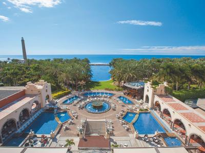 Vista aérea de una piscina de hotel de lujo con vista al mar y palmeras al fondo.