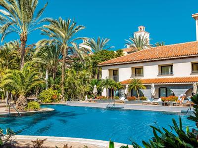 Hotel con gran piscina, palmeras y plantas florecientes bajo cielo azul claro.