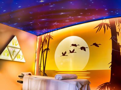 Chambre de bien-être chaleureuse avec table de massage, art mural de coucher de soleil et oiseaux, ambiance apaisante.