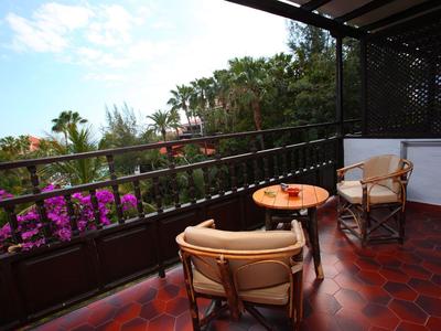 Balkon mit zwei Stühlen und Tisch, Blick auf blühende Pflanzen und Palmen.