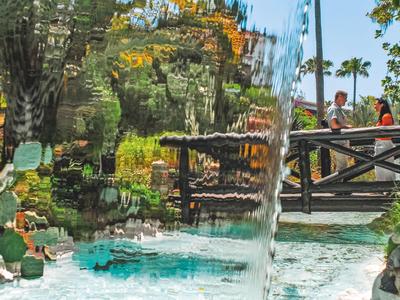 Fluss mit Wasserfall im tropischen Garten eines Hotels mit Holzbrücke und Sitzbereich.