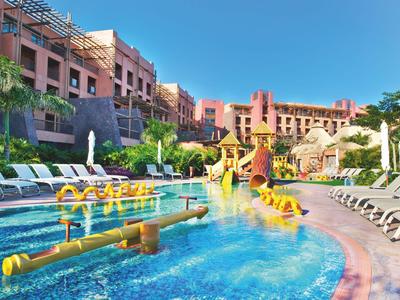 Hellblauer Pool mit Wasserspielplatz und Liegestühlen vor einem Hotelgebäude unter klarem Himmel.