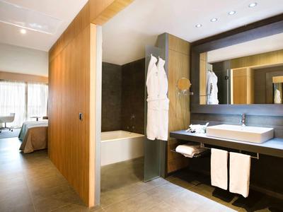 Baño moderno con bañera, lavabo, grandes espejos y vista al dormitorio.