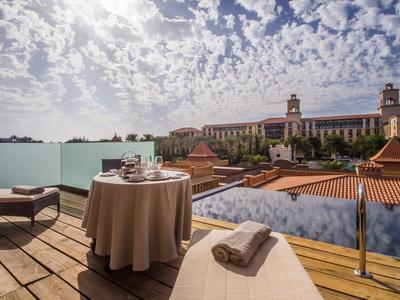Balcone con tavolo, sedia e lettino con vista sugli edifici dell'hotel sotto un cielo nuvoloso.