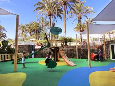 Kinder spielen auf einem bunten Spielplatz mit Rutsche und Schaukeln unter Palmen.