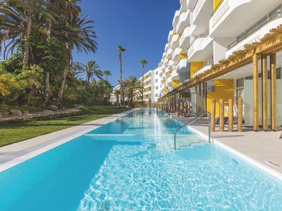 Moderner Außenpool mit klarem Wasser neben einem weißen Hotelgebäude und Palmen.