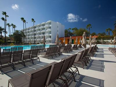 Hotelterras met ligstoelen en zwembad onder een blauwe hemel met palmbomen.