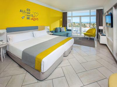 Licht ingericht hotelkamer met groot bed, gele accentmuur en zithoek met bank en stoel.