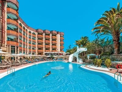 Hotel met groot buitenzwembad, parasols en palmbomen onder een heldere blauwe lucht.