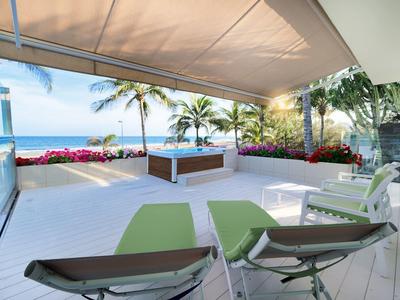 Bright hotel terrace with chairs, plants, and ocean view under a canopy.