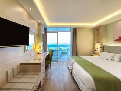 Modern hotel room with a large bed, wall-mounted TV, desk, and balcony with ocean view.