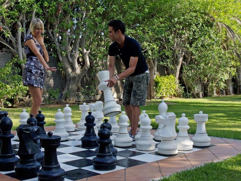 Two people playing chess on a large outdoor chessboard in a park-like garden.