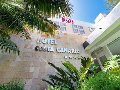 Hotel Costa Canaria building facade with palm tree and green plants under clear blue sky.