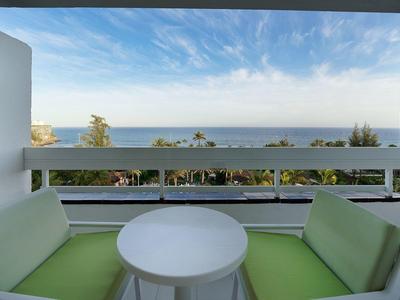 Two green cushioned chairs and a white round table on a balcony overlooking the sea.