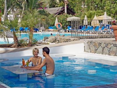 Couple relaxes in pool with built-in table, surrounded by lounge chairs and greenery.