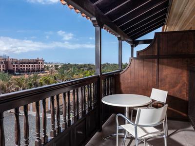 Balkon met een ronde tafel en twee stoelen met uitzicht op palmbomen en een gebouw in de verte onder een blauwe lucht.