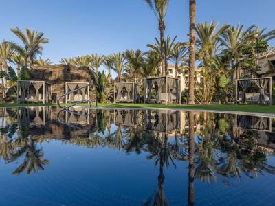Ein ruhiger Pool spiegelt Palmen und Bungalows unter klarem blauem Himmel.