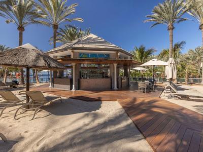 Strandbar met rieten daken omringd door palmbomen en ligstoelen onder blauwe lucht.