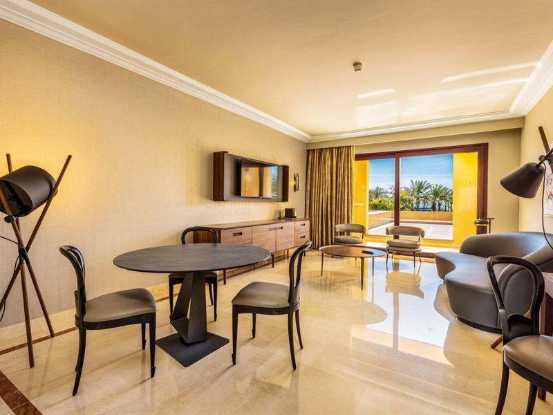 Ruime hotelkamer met eetgedeelte, bank, fauteuils en groot raam met zonnig uitzicht.