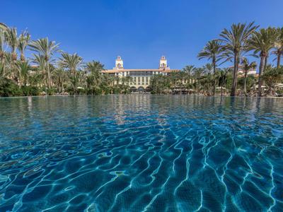 Großer Pool mit klarem Wasser und Palmen vor einem Hotel bei klarem Himmel