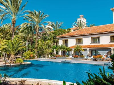 Resort met zwembad, palmbomen en een gebouw met twee verdiepingen onder een heldere blauwe lucht.